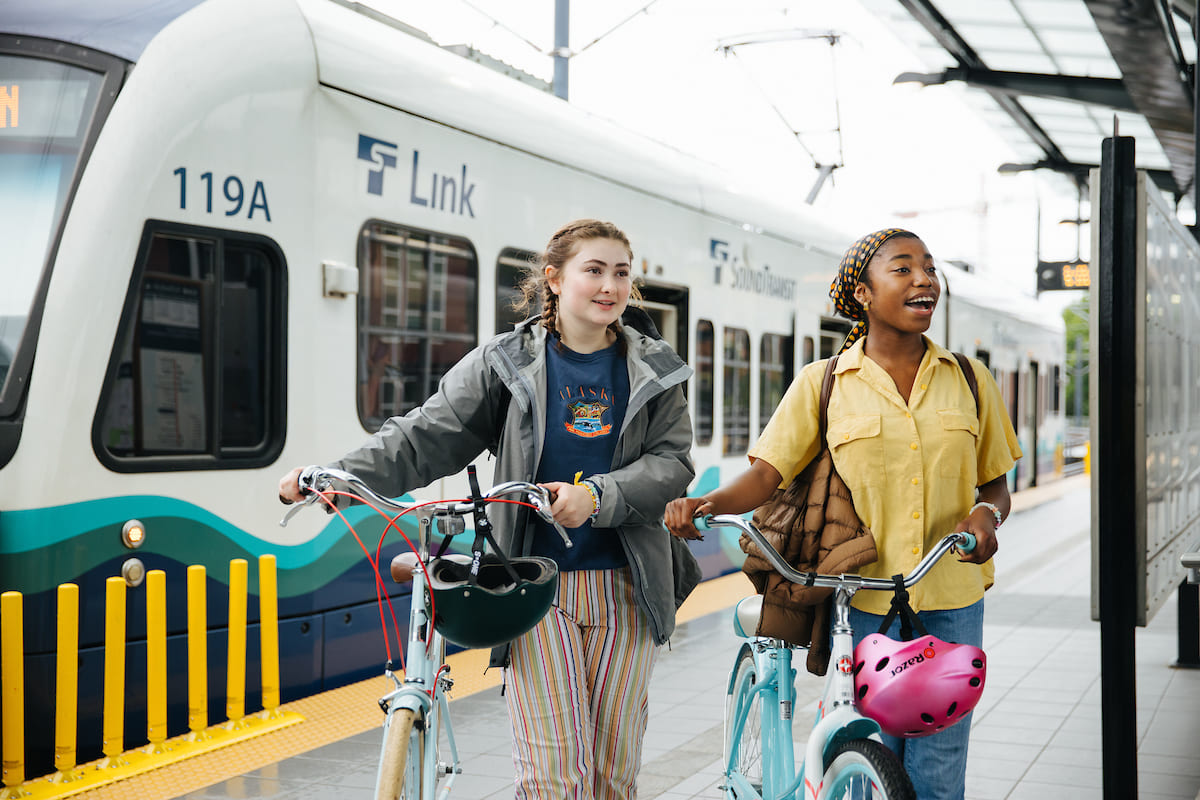 Dalawang teenager na may dalang bisikleta, na naglalakad sa Link light rail platform nang may tren sa background.  